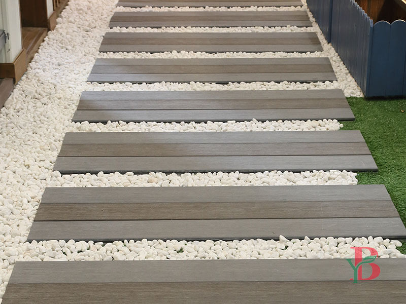 Horizontal grey-brown composite decking planks used as modern stepping stones in a garden landscape with white pebbles.
