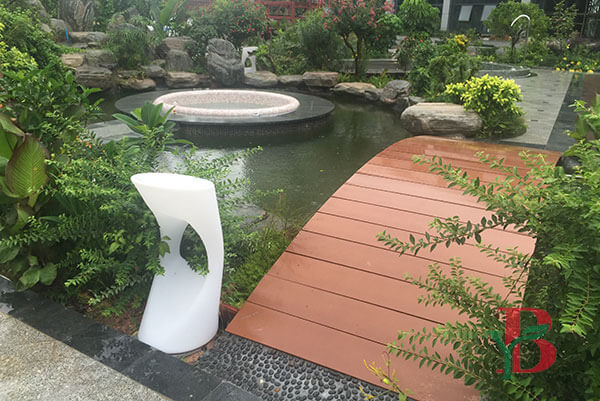 Side view of a curved red-brown composite decking bridge over a garden pond, showcasing durable WPC railings and high-quality wood grain texture by Bongywood.