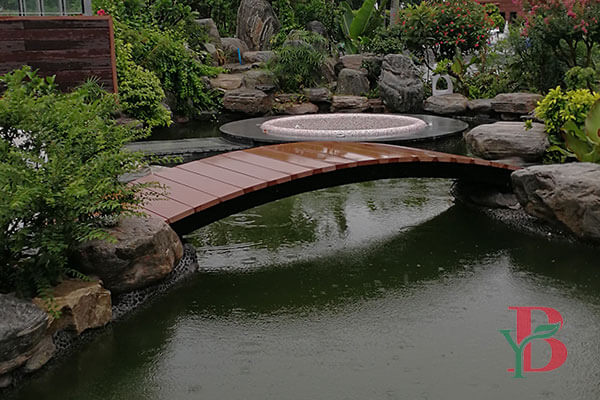 A tranquil garden landscape featuring a composite decking bridge leading towards a circular stone fire pit and modern sculptures, using Bongywood outdoor decking.