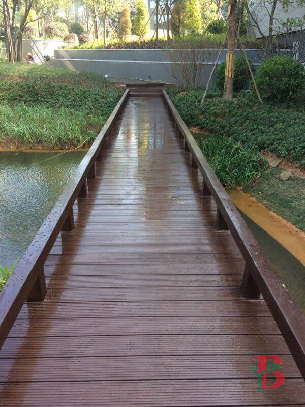 Dark brown water-resistant composite decking walkway built over a pond, featuring a durable grooved surface for slip resistance in garden landscapes.