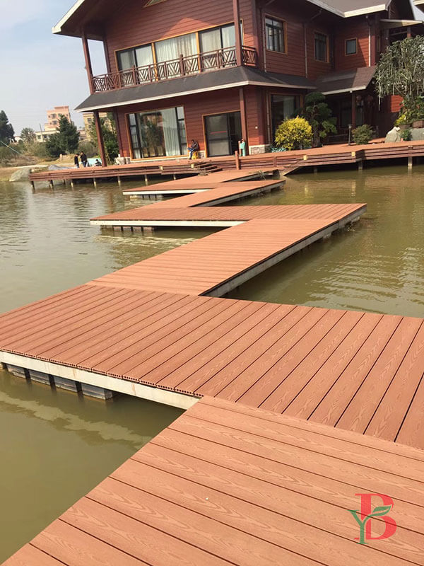 A zigzag-style floating composite decking path and pontoon on a lake, leading to a traditional house, showcasing Bongywood's orange-brown WPC boards.
