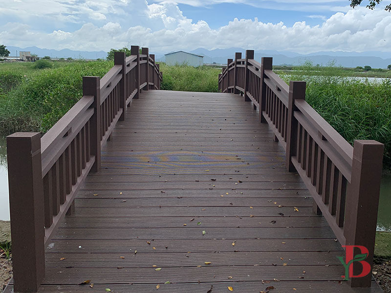 Sturdy arched bridge crossing a wetland, built with brown composite decking and matching railings.