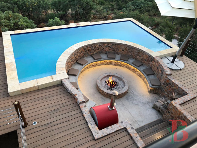 1.1bongywood-3d-embossed-composite-decking-fire-pit-pool Bongywood 3D embossed composite decking installed around a luxury curved swimming pool and outdoor fire pit area, showcasing natural wood grain texture and high slip resistance.